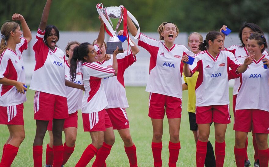 A women's football team celebrate holding a large trophy aloft. 