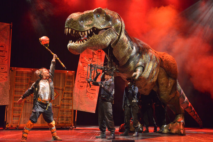 Large T-rex puppet being controlled by two puppeteers and another actor interacting with the dinosaur on stage