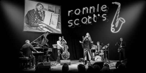 Black and white image of a band on stage with a neon sign of a saxophone and 'Ronnie scott's'