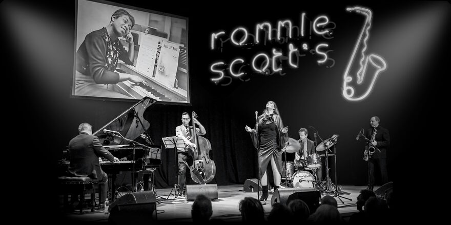 Black and white image of a band on stage with a neon sign of a saxophone and 'Ronnie scott's'