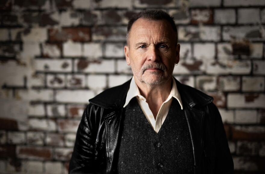 A portrait of Martin Simpson in a black leather jacket and waistcoat, standing against a textured brick wall.