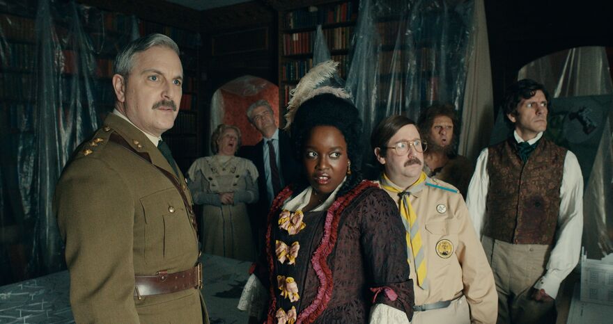 A group of people in various period dress stand in a library, covered in dust sheets, looking out of a window.  