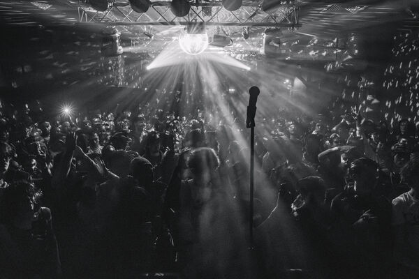 A black and white photo of a lively, dense crowd in a smoky music venue, illuminated by bright beams from a disco ball, with a silhouetted microphone stand in the foreground.