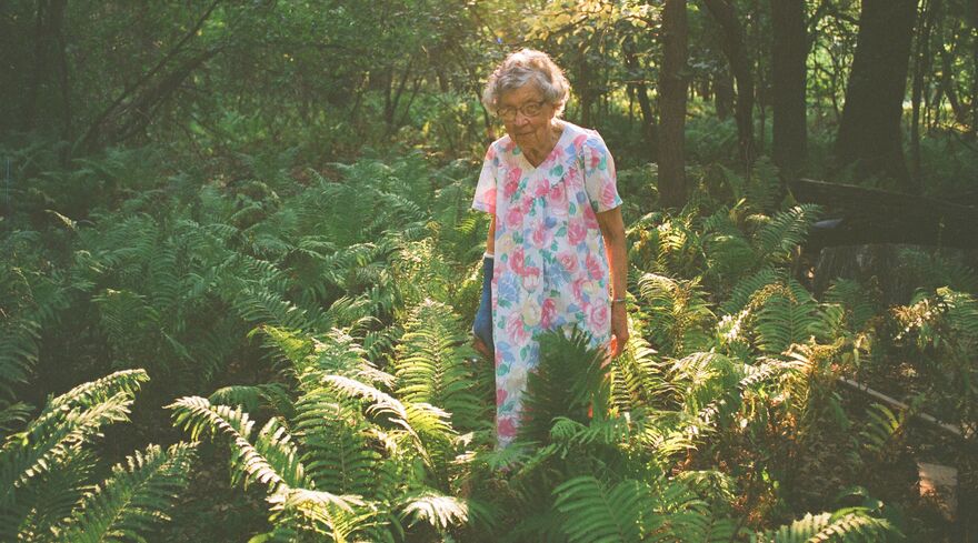 An older woman wearing a floral dress stands in the middle of a green woodland clearing. 