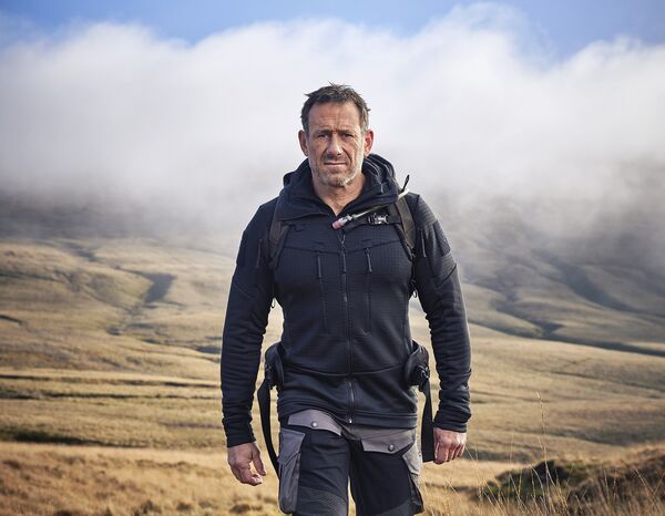 Jason Fox walking on grassy hills with cloudy blue sky behind 