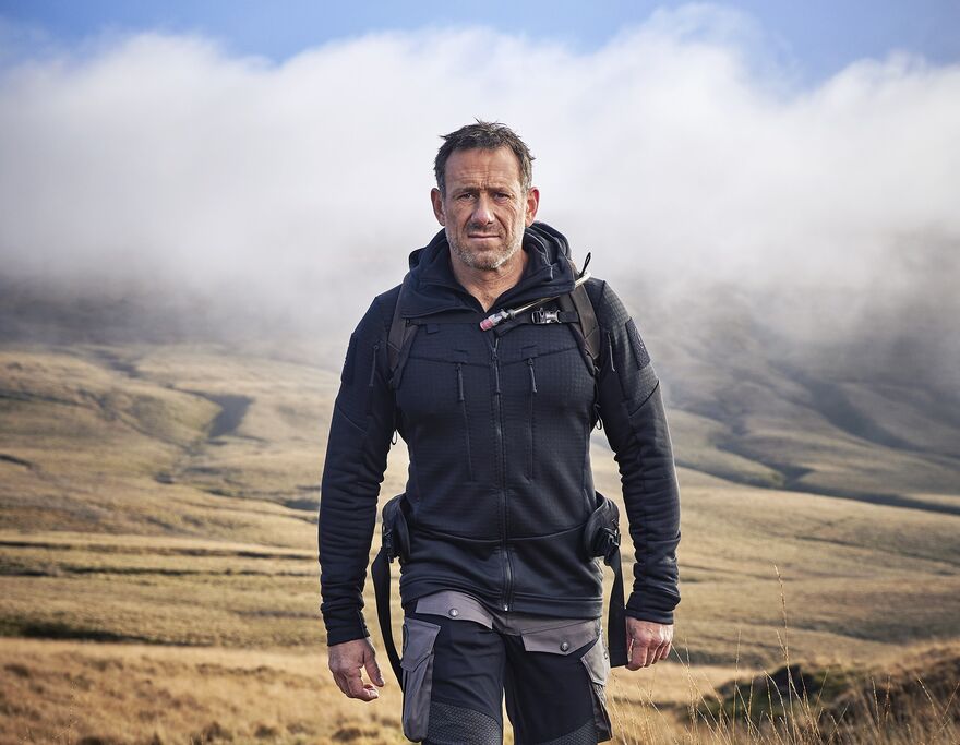 Jason Fox walking on grassy hills with cloudy blue sky behind 
