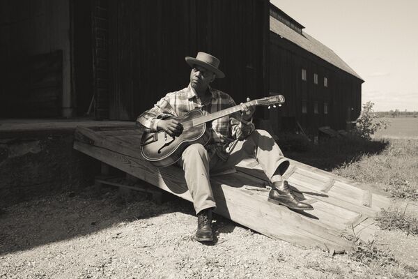 Eric Bibb sat on a ramp outside a building playing guitar, image is in black and white