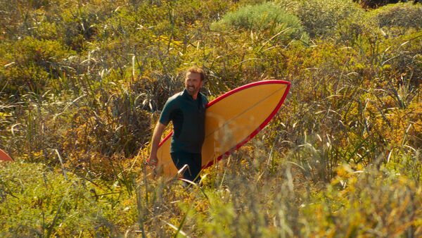 A man walks through tall grass with a surfboard under his arm. 
