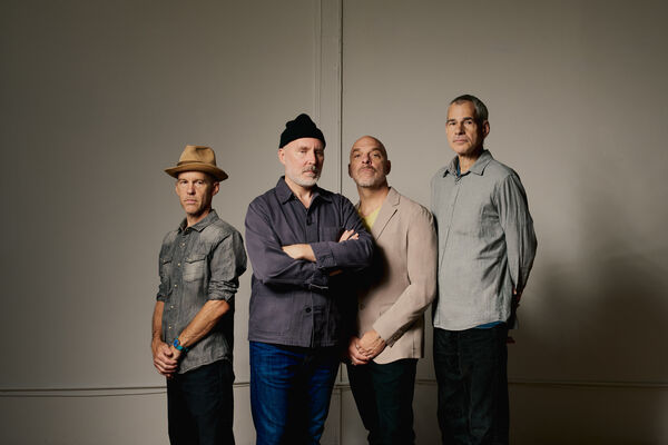 A studio portrait of the four members of the band The Bad Plus standing side-by-side against a plain, light-grey wall.