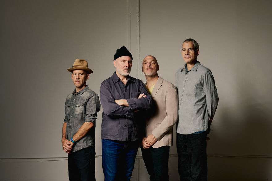 A studio portrait of the four members of the band The Bad Plus standing side-by-side against a plain, light-grey wall.