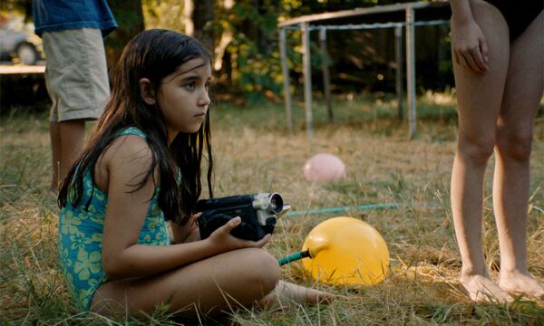 A young girl sits on the grass wearing a swimming costume holding a camcorder. 