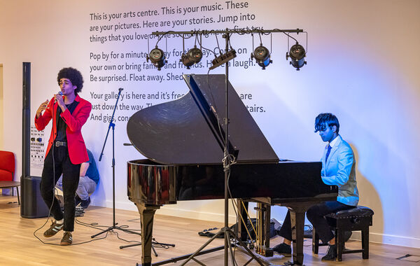 Two students perform in the Warwick Arts Centre foyer. One holds a microphone, the other plays the piano.