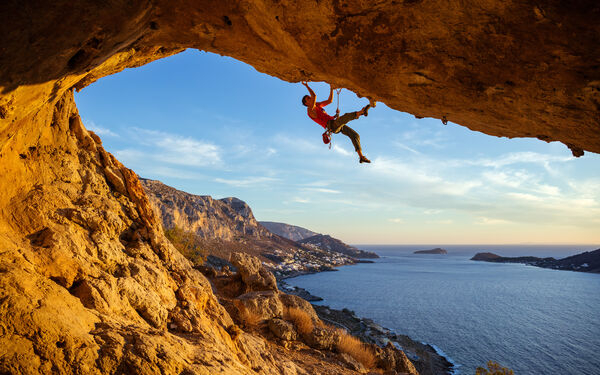 A rock climber scales the underside of a natural arch, with a panoramic view of the blue ocean and a coastal landscape in the background.
