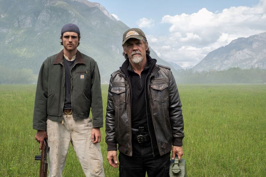 Two men, one older than the other, stand together looking to camera in an open green field. Mountains can be seen in the background. 