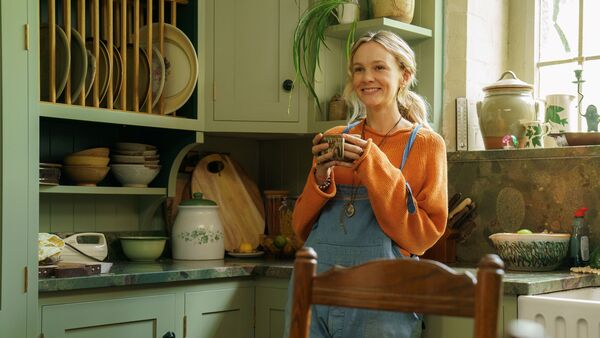 A woman in dungarees holds a mug leaning against a kitchen counter. 