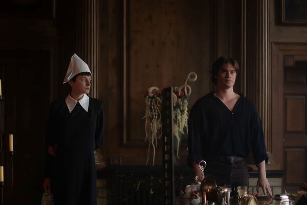 A man and woman dressed in all black clothing stand apart in a grand hall. 