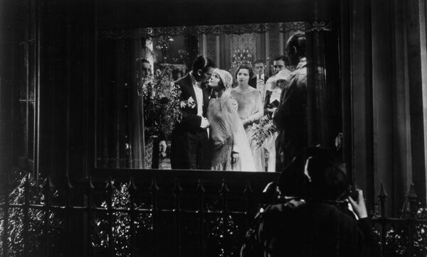 A man stood outside looks into a room through a window where poeple are dressed in fancy clothes. Image is in black and white