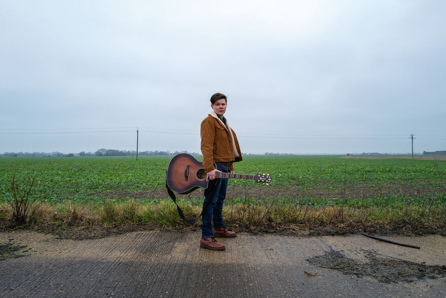 Singer Grace Petrie stands in the fields with a guitar.