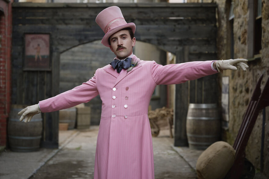 A man stands with his arms out wearing a pastel pink suit. 