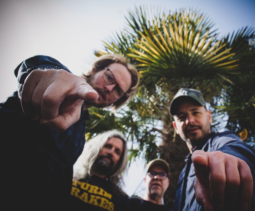 4 men standing in front of a palm tree with two pointing at the camera