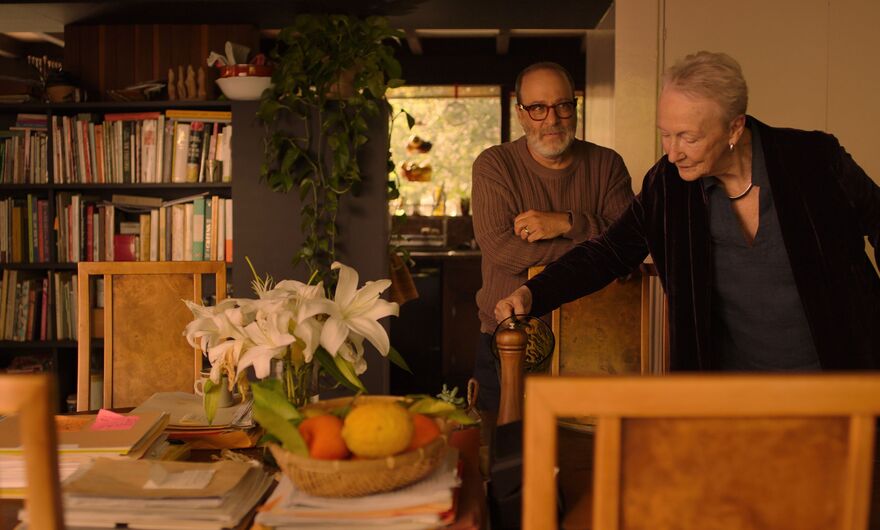 An older woman pours from a jug as a man stands watching beside her. They are stood in a dining room surrounded by books and piles of paper. 