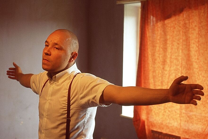 A man with a skinhead wears a white shirt with braces with his arms stretched wide. 