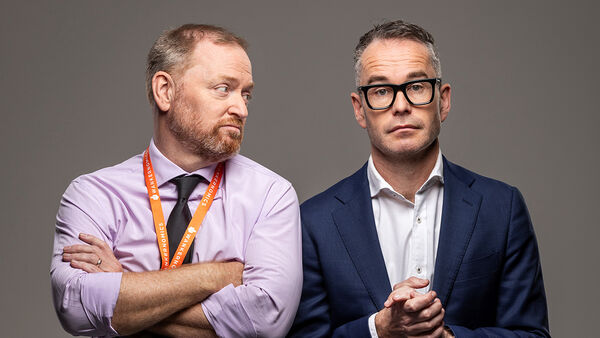 A promotional portrait of the corporate comedy duo, Wankernomics. Two men stand against a grey background in office attire, capturing their satirical corporate personas.