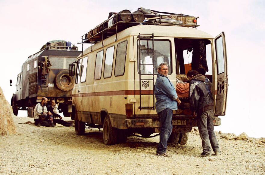 Two camper vans next to each other on a dusty road. Two men can be seen next to each vehicle. 