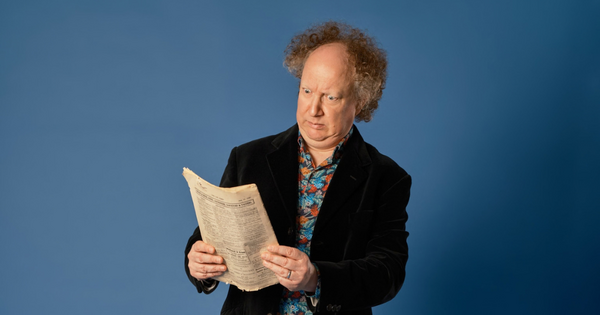 Comedian Andy Zaltzman holding and reading an old newspaper with a puzzled expression against a solid blue backdrop.