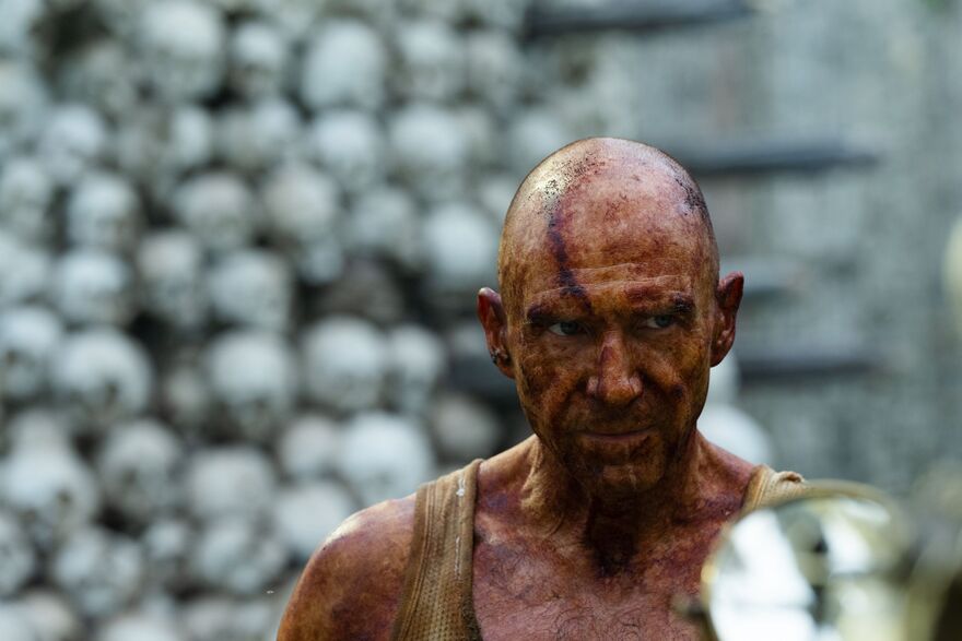 A man covered in dirt and blood. A stack of skulls can be seen in the background.