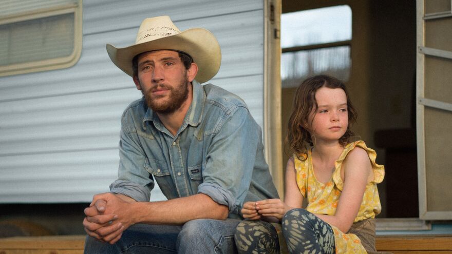 A man wearing denim and a cream coloured Stetson sits next to a young girl outside a trailer. 