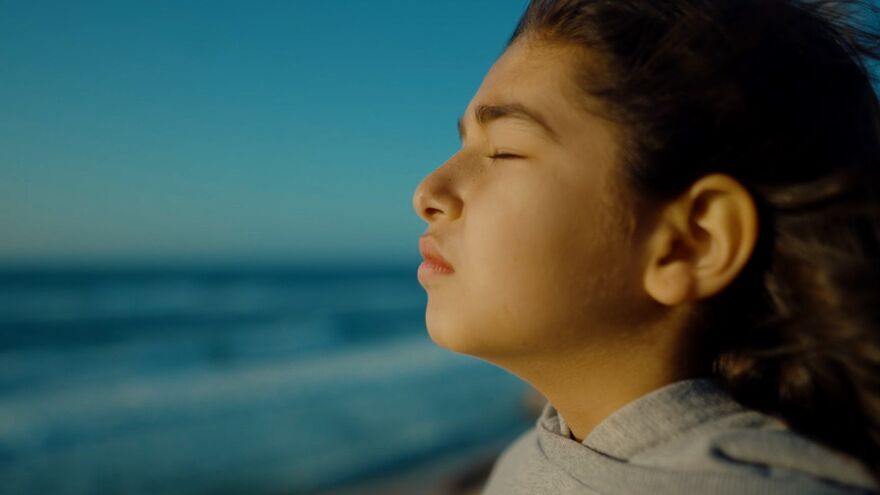 A young child stands near the shore with their eyes closed. 
