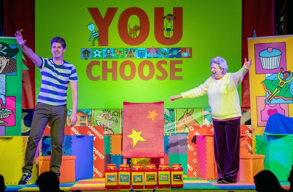 Two performers on stage with a starry chair between, 'You Choose' written in the background