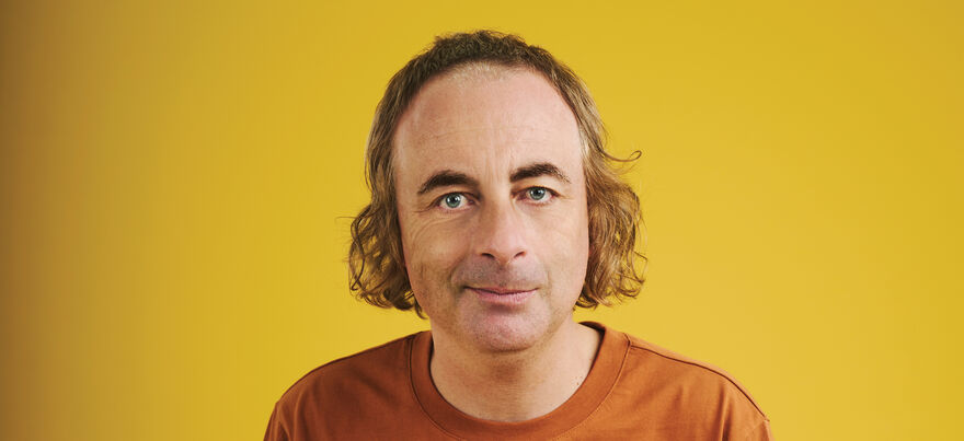 Paul Foot smiles at the camera in front of a mustard yellow background