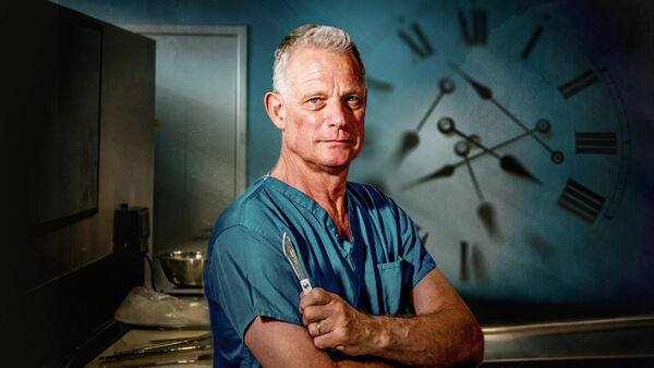Dr Richard Shepherd in scrubs with a large clock on wall with multiple hands