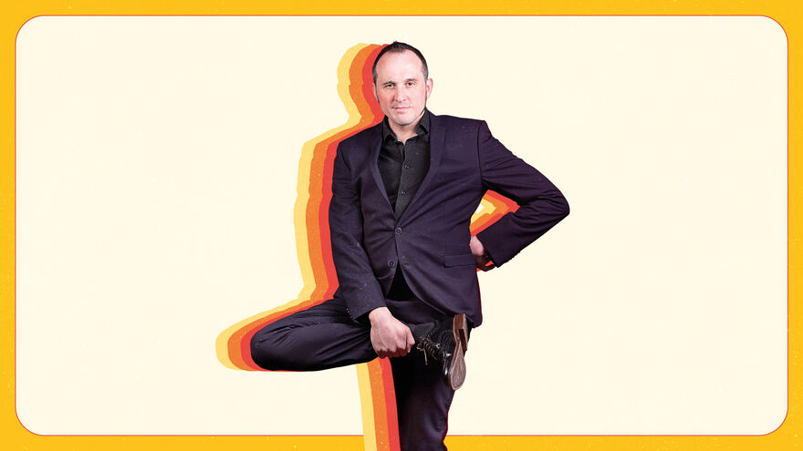 Phil stands on one leg, looking at the camera, with a pale yellow background
