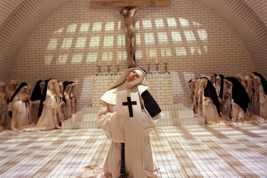 A nun stands at an awkward angle looking up at a crucifix. A large group of nuns are praying in the background. 