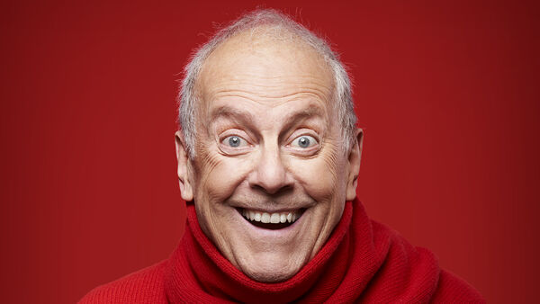 Giles Brandreth Smiling in a red turtleneck with a red background 