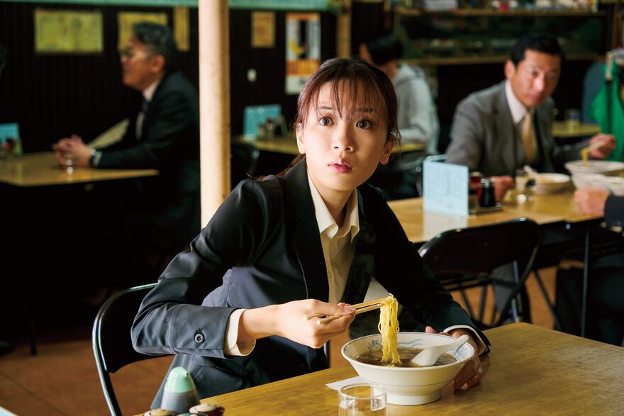 A young Japanese woman looks up startled whilst eating noodles.