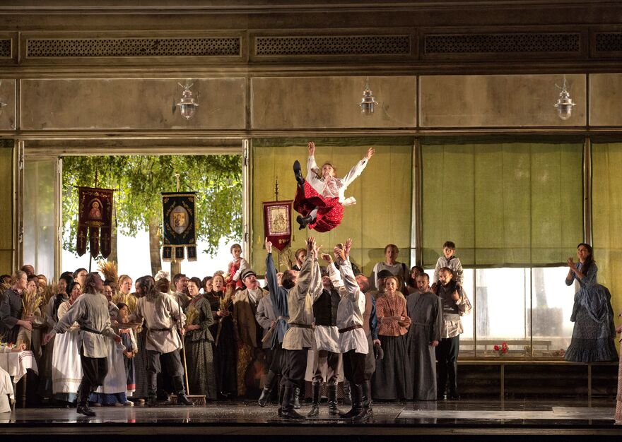A group of men throw a woman into the air. A large ensemble can be seen in the background.