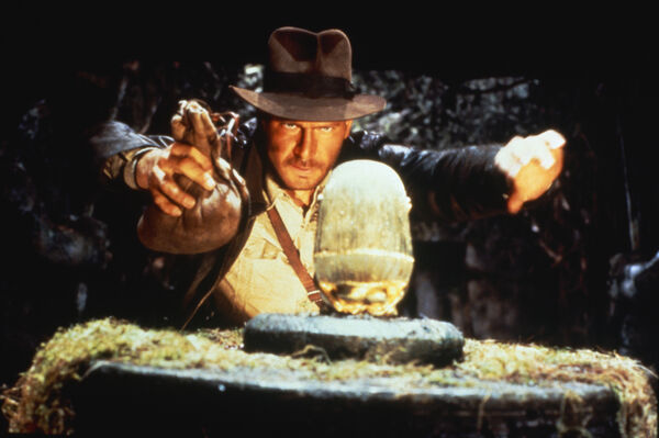 Harrison Ford as Indiana Jones in a cave in front of a golden object. He holds a brown bag. 