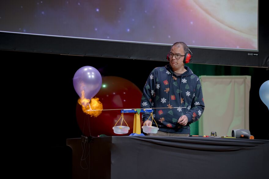 Man in a christmas jumper behind a scale with a stick on fire below a balloon
