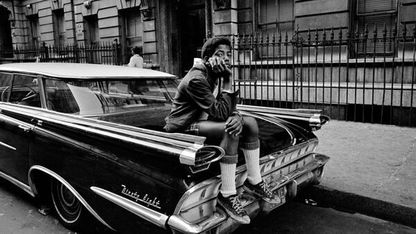 A black and white image of a young boy sat on the boot of a car. He looks to camera resting his hand on his cheek. 