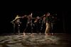 Six barefoot dancers in casual, dark clothing lunging forward dynamically on a stage lit with patterned floor lighting.