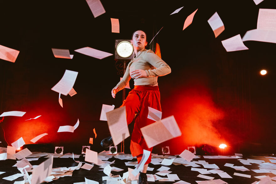 A performer from RPM kicks dynamically amidst a flurry of falling paper on a stage illuminated by dramatic red lighting.