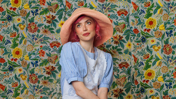 Tatty Macleod in a blue pintsripe top and a large pink hat, floral flower wallpaper in the background