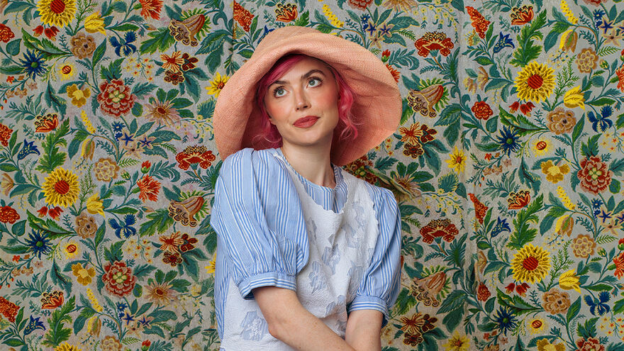 Tatty Macleod in a blue pintsripe top and a large pink hat, floral flower wallpaper in the background