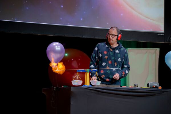 A presenter watches a purple balloon burst into flames during a science experiment on stage.