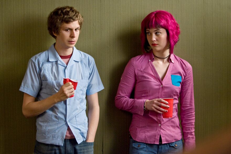 A teenage boy stands anxiously next to a teenage girl with bright pink hair. 