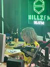 Kate Hills sitting in a radio studio at a desk with broadcasting equipment, in front of a glowing green neon sign that reads 'HILLZFM 98.6FM'.